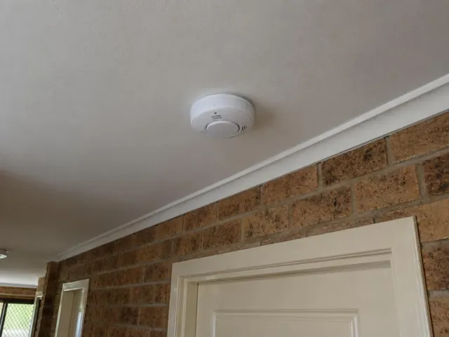 Ceiling-mounted smoke alarm above a brick wall and interior door in a home in Worongary